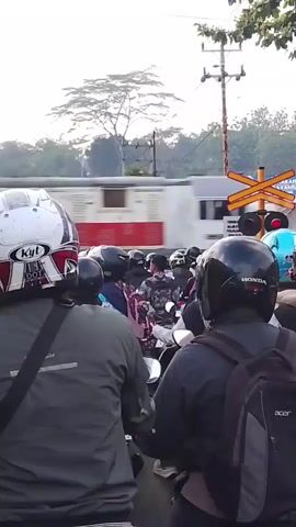 The video shows a congested traffic scene at a railway crossing. A train is passing through the crossing from left to right. Several motorcycles are stopped in traffic, waiting for the train to pass. Most people are wearing helmets, and some are carrying backpacks. The weather appears to be clear.