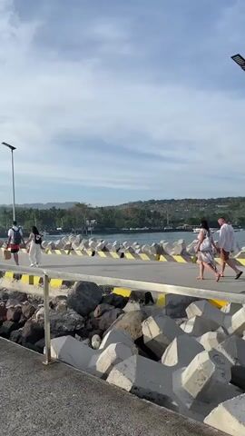 A group of people are walking along a concrete pier. The pier is lined with rocks and concrete barriers on one side, and a yellow and black railing on the other. In the background, there is water, boats, and a coastline with trees and buildings. The sky is cloudy. The people are dressed in casual clothing and some are carrying bags. The scene appears to be a public walkway near a harbor or port.