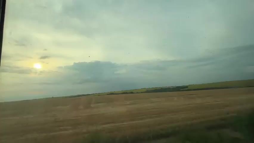 The video shows a view from inside a vehicle, likely a train or a bus, looking out a window. The landscape outside is a rural scene with fields of harvested wheat and some areas of crops, possibly sunflowers, in the distance. The sky is overcast with dramatic clouds, and the sun is partially visible, casting a soft light. There are trees and vegetation in the foreground and along the horizon. The camera is stationary, and the video captures the subtle changes in light and cloud movement.