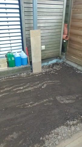 The video shows an area under construction or renovation. A yellow plate compactor is sitting on a patch of dirt and gravel. Adjacent to this is a building with closed white blinds and light brown wood paneling. Near the building are containers of blue and green liquid. The surrounding area is mostly dirt and gravel with some construction materials like wooden planks, metal pieces, and a large white bag of Steffes stone. Across the street, there is a building with white walls and a blue fence. Green trees are visible in the background.