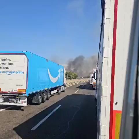 The video shows a traffic jam on a highway, with several trucks stopped on the road. Thick plumes of black smoke are visible in the distance, rising from an area off to the side of the highway. The smoke suggests a fire is burning, and the trucks are likely stopped due to the fire or related road closures. The sky is clear and blue, and the surrounding landscape appears to be dry and grassy, with some trees visible. The camera appears to be inside one of the vehicles, looking out at the scene.