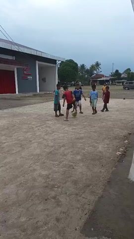 A group of children are playing soccer on a dirt field in front of a building with red doors and a white facade. The sky is overcast. The children are wearing casual clothes. The ball is yellow.