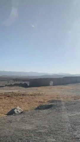 The video depicts a view from a moving vehicle, possibly a car or truck, driving along a dirt road. The landscape is barren and dusty, with low-lying buildings on the side of the road. In one shot, there is a building with graffiti written in red, partially obscured. Mountains can be seen in the distance under a clear sky. There are no people visible in the video.