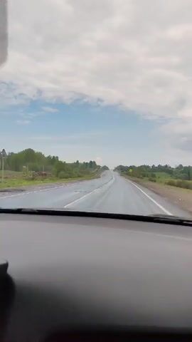 The video shows a view from inside a car driving on a wet road. The road is straight with two lanes in each direction, separated by a median. There are trees and bushes along the sides of the road. The sky is cloudy, with patches of blue. The car is moving forward, and the windshield wipers are on. The dashboard of the car is visible.  The road surface reflects the sky. There are guardrails on the sides of the road. Road signs are present.