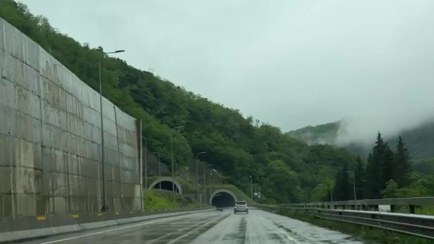 The video shows a car driving towards a tunnel on a highway.  The road is wet and it appears to be raining. On the right side of the road are mountains covered with green trees and some fog.  The fog mostly obscures the upper portion of the mountain.  The highway is flanked by a gray concrete barrier wall on the left. Streetlights are visible on both sides of the road.