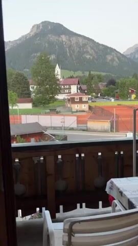 The video shows a view from a balcony. In the background is a mountain with a town nestled at its base. A church steeple is visible among the buildings. In the foreground, the balcony has a wooden railing and a table with a white tablecloth is partially visible. There is a tennis court to the right. The sky is light blue. The camera is mostly still.