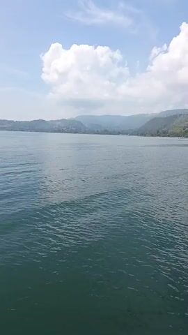 A wide shot of a large body of water, likely a lake or ocean, under a bright, partly cloudy sky. The water is a deep blue-green, with small ripples on the surface. In the distance, a lush green coastline is visible, with hills and mountains forming the backdrop. The scene appears peaceful and serene, with no visible boats or people.