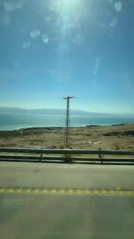 The view from a moving vehicle shows a coastal landscape under a bright, sunny sky. A highway with a grey guardrail and yellow-striped lines in the foreground runs parallel to the shore. The shoreline features a beach with light-colored sand or earth that fades into a body of water, possibly a sea or large lake, with a turquoise hue. In the distance, a hazy outline of land or mountains is visible on the horizon. A utility pole with wires stretches across the scene, connecting the land to the electrical grid. The atmosphere appears clear, but there are some reflections and minor visual artifacts on the windshield of the moving vehicle.