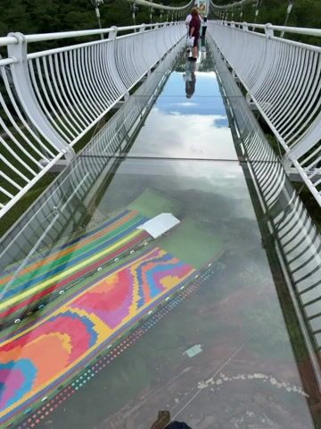 This video shows a person walking across a glass-bottomed suspension bridge. The bridge has white metal railings and is suspended high above a lush green forest. Below the glass floor, a colorful, geometric pattern is visible on the ground. The sky above is a mix of blue and white clouds. The camera pans and tilts, offering different perspectives of the bridge and the surrounding landscape. The person walks slowly, pausing to look down at the view. The video captures the sense of height and the unique experience of walking on a transparent bridge.