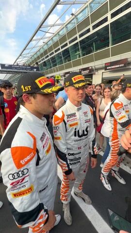 This image captures two male racing drivers, likely winners, being interviewed on what appears to be a pit lane or grid area at a racetrack. Both drivers wear white and orange racing suits adorned with sponsor logos such as "TREESOR," "Audi Sport Customer Racing," "freem," "IRELLI," "GT WORLD CHALLENGE," "PST GRUPPEN," and "AVISTA." They are also wearing matching black baseball caps featuring a gold laurel wreath design and "1ST" embroidered prominently in yellow, signifying their victory.

The driver on the left is actively speaking, looking towards the interviewer or his teammate with a slight smile. The driver on the right is listening attentively, also smiling. An unseen interviewer holds a black microphone, branded with "GT WORLD CHALLENGE," towards them.

In the background, a crowd of spectators and crew members can be seen, some smiling and observing the interview. Behind them are modern, multi-story buildings with numerous windows, suggesting pit garages or a hospitality complex. One door is labeled "SCRUTINEERING GT3." High above, a gantry structure spans the area, displaying the text "AUTODROMO NAZIONALE MONZA," confirming the location as Monza, Italy. An Italian flag is visible fluttering in the distance against a partly cloudy blue sky. The overall atmosphere appears celebratory and energetic, suggesting a post-race event during daytime with pleasant weather.