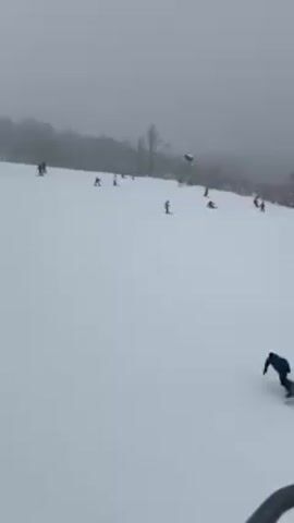 The video shows a snowy hillside with several people skiing and snowboarding down it. The weather is overcast and foggy.  The trees bordering the hill are covered in snow.  A skier in a blue jacket and red pants is visible in the foreground. Other skiers and snowboarders are spread across the slope.  One person is sitting on the snow.