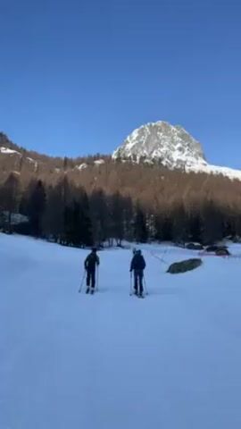 Two people wearing backpacks and skis walk across a snow-covered field. The field is flat and groomed. There are trees bordering one side of the field. In the background, a mountain towers over the scene. The sky is clear and blue.  The camera follows the two people as they move.