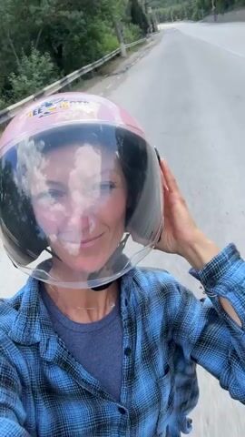 A woman wearing a blue and black plaid shirt and a pink helmet with a clear visor rides on a scooter or motorcycle. She is holding the visor with her right hand and smiling at the camera. The background shows a road with trees and power lines on either side, suggesting a rural or suburban setting. The sky is partly cloudy. The video appears to be a selfie taken by the woman as she travels.