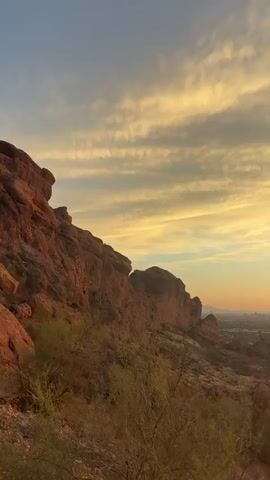 The video begins by showing a rocky, mountainous terrain covered in sparse desert vegetation. As the camera pans to the right, a panoramic view of a city unfolds in the distance, nestled between rolling hills. The sky is ablaze with the vibrant colors of a sunset, with streaks of orange, yellow, and pink clouds painting the horizon. The sun, a bright orb of light, is slowly descending, casting a warm glow over the landscape. The video captures the tranquil beauty of nature as day transitions into night.