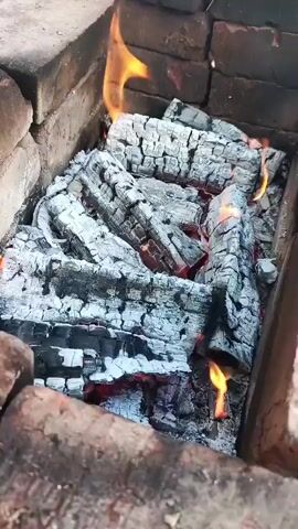 A close-up overhead shot of burning logs in a brick pit. The logs are mostly ash-covered, with some areas still glowing red and small flames licking the sides of the wood. The pit appears to be made of rough, reddish-brown bricks. The lighting is dim, focusing on the fire.