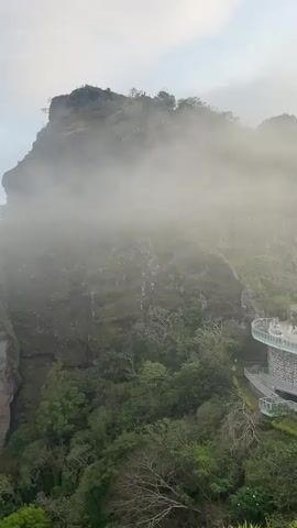 The video pans from a mountain shrouded in mist to a view of a valley below. The mountain is covered in green vegetation and has a rocky cliff face. The mist obscures the view of the top of the mountain in the beginning of the video, but it gradually dissipates as the camera pans to the right. A white, multi-level structure with circular balconies is visible on the right side of the mountain. The valley below is filled with trees and a small town. The sky is overcast with a light blue hue.