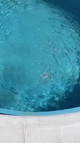 A video shows a swimming pool from above. The water is a clear blue, and there are ripples on the surface. The pool is surrounded by a tiled deck. In the background, there are lounge chairs and a fence. The video also shows a ladder entering the pool. The camera pans slightly to the left, showing more of the pool and the surrounding area.