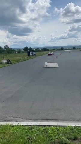 A red Tesla Model 3 is seen driving on a paved track, possibly a race track or a closed course. The car is red and appears to be the only vehicle actively moving on the track in the immediate vicinity. The car is making a turn. In the background, there is a large inflatable building with the words "ТРЕНИНГ-ЦЕНТР" and "АСАМАРА" written on it. There are also other cars parked in the distance near the inflatable building. The track is lined with orange cones, suggesting it is set up for some kind of driving event or training. There are also some white barriers on the track. The sky is cloudy, with patches of blue visible. The surrounding environment appears to be rural or semi-rural, with grassy areas and distant hills. The video is shot from a stationary position beside the track, looking towards the car.