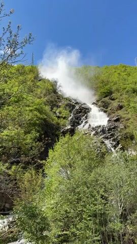 The video shows a waterfall cascading down a rocky slope. Lush green trees surround the waterfall, growing on the hillside. The water flow is strong, creating a mist that rises into the air. The sky is blue, and there are no people visible in the scene. An electrical tower can be seen among the trees at the top left of the frame.