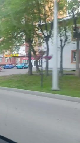 The video pans across a street scene in front of a multi-story building. Several cars are parked along the street, including a red SUV, a blue sedan, and a white car. People are visible walking and standing near the cars and on the sidewalk. The building has a mix of brick and white facade with many windows. There are trees and green spaces along the street. The sky is visible with some clouds. The camera appears to be in motion, suggesting the video was taken from a moving vehicle.