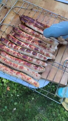 A close-up overhead shot shows a person's hands using tongs to turn sausages on a grill. The sausages are browning and appear to be nearly cooked. The grill is set on a wooden surface outdoors, with green grass visible in the background. A water bottle and other items are also present in the scene. The video then cuts to a shot of the sausages being placed into a metal pot.