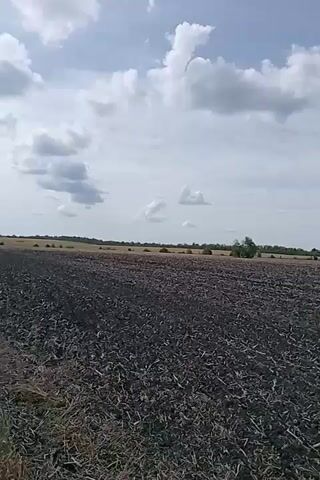 The video shows a wide shot of a freshly plowed field under a partly cloudy sky. The field is dark brown and appears to be recently tilled, with remnants of crops visible. In the distance, a line of trees marks the horizon. The camera pans slowly across the field, capturing the vastness of the agricultural landscape. The overall impression is one of open space and the preparation of land for planting. There are no people or animals visible in the video. The lighting suggests it is daytime, with sunlight filtering through the clouds. The scene is tranquil and rural.