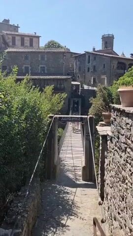 The video shows a sunny day in what appears to be a historic European village. A pedestrian suspension bridge made of wood and metal spans a narrow gorge. On the bridge, two people, one in light-colored clothing and another with a lighter backpack, are walking away from the camera towards a stone building with a tower in the background. The bridge is framed by lush green trees on the left and a stone wall on the right. The buildings in the background are constructed of stone and have tiled roofs, typical of old European architecture. The sky is clear and blue, indicating good weather.