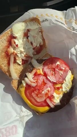 The video shows a close-up of a McDonald's burger wrapped in branded paper. The burger appears to be made with a thick bun, a beef patty, cheese, tomatoes, onions, and sauce. A hand is shown assembling the burger and then holding it as it is being taken out of the wrapping. The lighting suggests the video was taken outdoors in daylight.