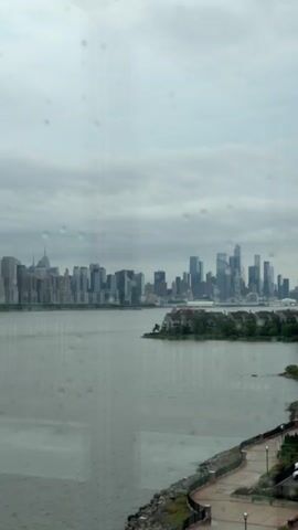 The video captures a wide shot of a city skyline across a body of water. The sky is overcast, and there are visible raindrops on the lens, suggesting it is raining. In the foreground, a paved walkway with lampposts follows the edge of the water, bordered by rocks and some greenery. Across the water, a dense cluster of skyscrapers forms the city skyline, with some lower buildings and a small island with houses in between the foreground and the main skyline. The water appears calm, reflecting the muted tones of the sky and buildings. A few cars are visible parked near a modern building on the right side of the frame.