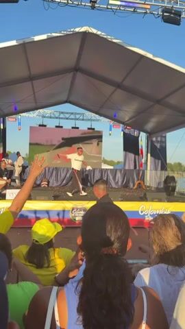 The video shows a crowd gathered to watch a performance on an outdoor stage. The stage has a large screen displaying a looping image or video. A male performer, dressed in a white t-shirt and black pants, is actively singing and gesturing on the stage. The crowd is a mix of genders and ages, many wearing yellow clothing and hats. The stage is adorned with Colombian flags. The overall atmosphere appears to be a lively outdoor festival or concert.