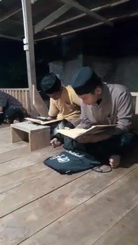 Several young men are sitting on a wooden porch, studying books. All are wearing similar black caps and are seated on the floor with their legs crossed. In front of each person is a small wooden stool upon which their books are placed. The porch has a simple roof structure with wooden beams, and the scene appears to be at night or in low light. The background shows a dark, indistinct view of the outdoors. Some have their bare feet visible.