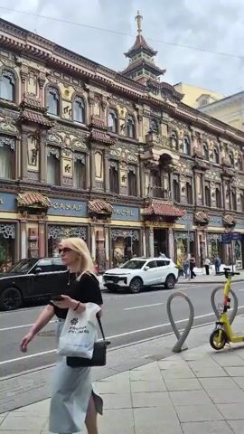 The video shows a street scene in front of a building with ornate, East Asian-inspired architecture. Cars are parked and driving on the street, and there are several electric scooters docked on the sidewalk. People are walking on the sidewalk, some appearing to be entering or exiting the building. The sky is overcast.