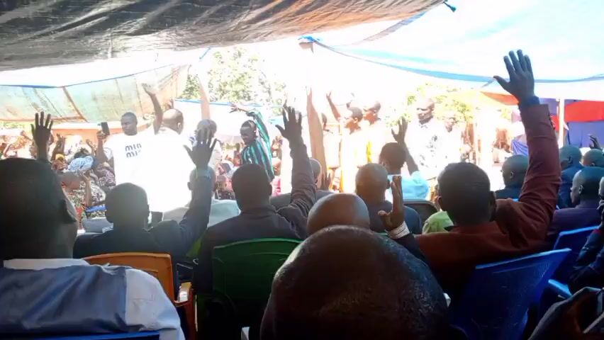 The video shows a gathering of people, mostly men, under a tent-like structure made of blue and white tarps. Many people are seated in plastic chairs, while some stand near the front. Most people have their hands raised in the air, appearing to participate in an event or ceremony. The crowd is predominantly Black, and clothing styles vary from casual to formal. The environment appears to be outdoors, with trees and buildings visible in the background. The audio suggests some type of sermon, with the speaker’s voice echoing throughout.
