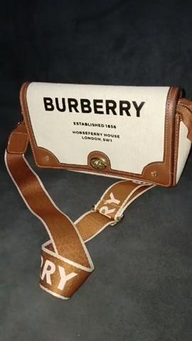 The video shows a Burberry shoulder bag with a white canvas body and brown leather trim. The front of the bag displays the Burberry logo in black, along with "ESTABLISHED 1856" and "HORSEFERRY HOUSE LONDON, SW1" also in black. The bag has a brown and white strap with the Burberry logo printed on it. The background is a dark, textured surface.