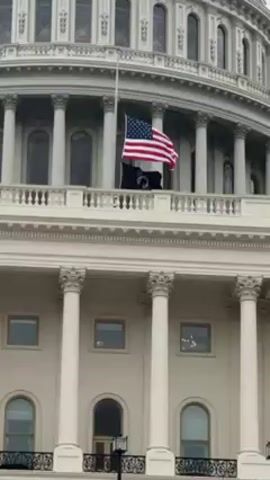 The video shows the United States Capitol Building on an overcast day. The American flag is at half-staff on the dome of the building. A black flag with a logo of a white eagle is hanging below the American flag. The dome of the building is white and has many windows and columns. The building appears to be made of stone.