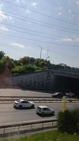 The video shows a view from a moving vehicle, likely a car, looking outwards towards a highway and its surroundings. On the highway, several cars are visible, including a dark-colored sedan, a white SUV, and a white truck. The highway appears to be elevated in some sections, with concrete support structures and railings. Overhead, power lines and a large transmission tower are prominent against a bright sky with scattered clouds. The roadside features lush green vegetation, including trees and bushes, and some areas of rougher terrain. A curved road sign with red and white markings is visible on the left side of the road. The perspective suggests the vehicle is traveling alongside or under an overpass. The overall scene depicts a typical urban or suburban highway environment on a sunny day.