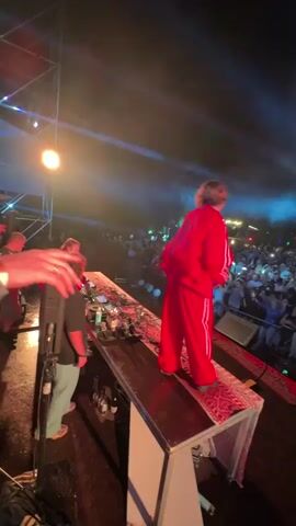 A person in a red tracksuit stands on a raised platform covered with a rug, facing away from the camera towards a crowd at an outdoor nighttime event. They are positioned to the right of a long table laden with electronic DJ equipment and bottles. To the left of the table, another person wearing a black t-shirt and light-colored pants, also facing away from the camera, appears to be operating the DJ equipment. Bright stage lights, including a spotlight and a hazy blue light, illuminate the scene. The crowd, visible in the background, is densely packed and illuminated by the stage lights. The overall environment suggests a concert or festival setting.