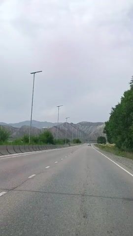 A vertical video shows a highway in a mountainous area under a cloudy sky. The road is asphalt with white lane markings and a concrete barrier on the left side. Several tall lamp posts are visible along the road. In the distance, a white car is traveling on the highway, and mountains with sparse vegetation form the backdrop. Trees are present on the right side of the road. The video appears to be filmed from a moving vehicle.