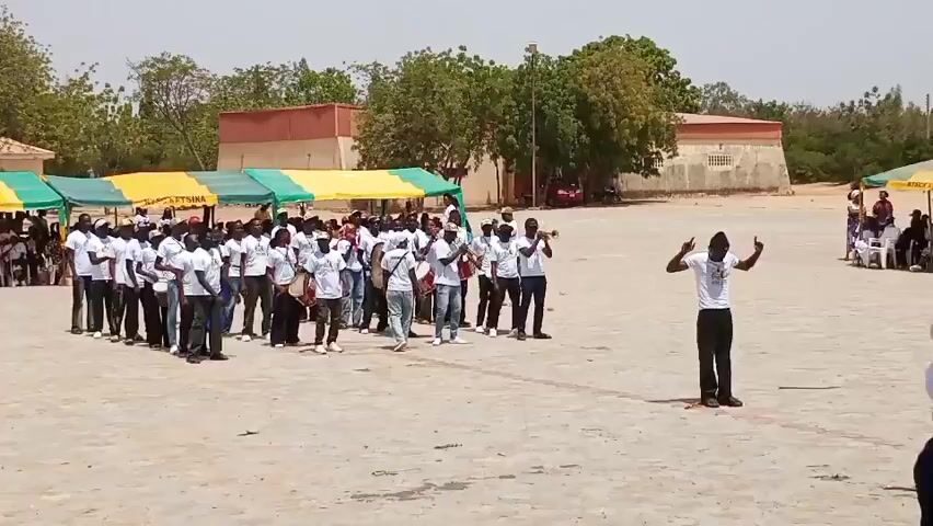 A group of people in white t-shirts and black pants are gathered in an outdoor, open area. Some are holding musical instruments like drums and trumpets, suggesting they are a marching band. In the foreground, a person in a white t-shirt and black pants, possibly a conductor, is facing the group and making hand gestures. There are tents set up in the background, and some spectators are seated on chairs. The environment appears to be a sunny day.