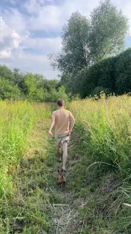 A person is walking on a dirt path through tall grass with a small dog following behind. The person is wearing a tan t-shirt and tan shorts. The dog is brown and appears to be small. The path leads towards a body of water and is lined with lush green vegetation. Trees and a cloudy sky are visible in the background.