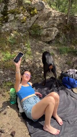 A long shot shows a woman sitting on a black blanket with two dogs, one black and one brown. The woman wears a blue top and denim shorts, glasses, and has a phone in her hands. The black dog wears a pink collar, and the brown dog scurries towards the water. The scene takes place outdoors near a lake, with trees and rocks in the background.