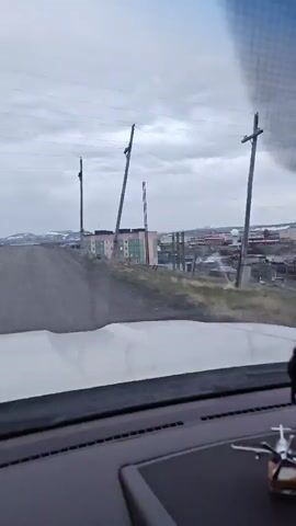 A video taken from inside a vehicle shows a desolate, cloudy landscape. A dirt road stretches ahead, leading towards a small town with several multi-story buildings and scattered houses. Tall utility poles with wires are visible along the road and throughout the town. The buildings in the town are brightly colored, with sections of pink, green, and blue. One tall, slender tower with a ladder or cage structure runs up its side is also prominent in the middle ground. The overall environment appears to be a remote, possibly northern, location with a stark and overcast atmosphere.