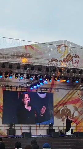 A woman in a black jacket and long white skirt stands on a stage, singing into a microphone. She is visible in real life and also in a large video screen on the left side of the stage. The stage is large and appears to be outdoors. It has an overhang and is decorated with lights. Behind the stage is an image on the back wall with a man and woman in what appears to be military uniforms. On the top right of the backdrop is the date "9 мая" [May 9th] and the words "День Победы" [Victory Day]. A crowd is gathered at the bottom of the screen, watching the performance. The sky is cloudy.