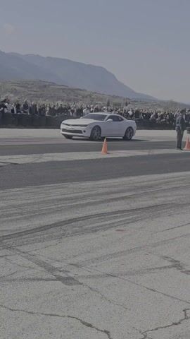 The video shows a drag racing event taking place on a paved track lined with spectators. In the first shot, a white Chevrolet Camaro is staged at the starting line, positioned to the left of an orange traffic cone. A crowd of people watches from behind a barrier on the left side of the frame, with mountains visible in the background. The second shot shows a gray BMW passing the Camaro, moving from left to right. The remaining shots primarily focus on the Camaro as it accelerates down the track, kicking up some smoke. The camera follows the car as it crosses the finish line, and the crowd of onlookers remains visible throughout the video.