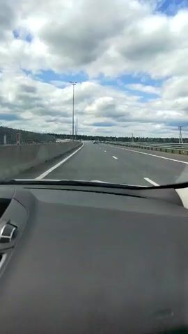 The video is taken from inside a car driving on a highway. The view is of the road ahead and the sky above. The road has multiple lanes and is bordered by a barrier on the left and guardrails on the right. There are streetlights and some trees visible in the distance. The sky is cloudy with patches of blue sky. Other cars can be seen on the highway in the distance. The dashboard of the car is visible in the foreground. The overall impression is that of a car journey on a highway on a cloudy day.