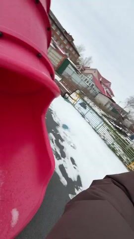 A woman is filming herself with a wide-angle lens while walking through a snowy outdoor playground area in Reshetnikovo, Russia. She is wearing a brown belted puffer coat and her hair is pulled back with a black claw clip. The ground is covered in snow, showing footprints and ski tracks. In the background, there are bare trees, various pieces of colorful playground equipment (including swings and a climbing structure), and residential apartment buildings. The sky is overcast, indicating a cloudy daytime. The woman walks slowly, looking around at her surroundings.