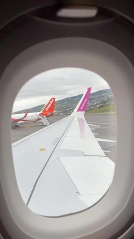 The video shows a view from an airplane window, focusing on the plane's wing. In the background, another airplane with the EasyJet logo is visible, with a staircase attached, presumably for boarding or deboarding passengers. The sky is overcast. The window frame is visible, giving context to the viewpoint. The general setting appears to be an airport runway or tarmac.