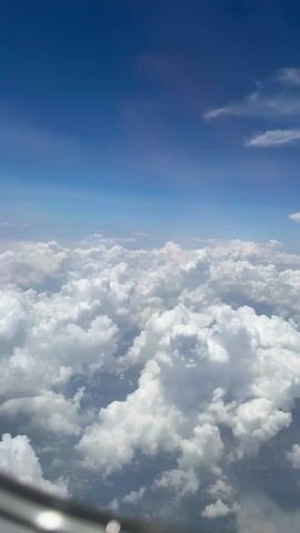 The video displays a view from an airplane window, showing a dense layer of white, fluffy clouds. Above the clouds, there's a clear blue sky. The edge of the airplane window is partially visible in the bottom left corner of the frame.