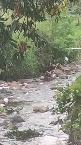 The video shows two people in a river full of trash. The river flows between rocks. One person is seated on a rock in the middle of the river, facing away from the camera. The second person is further up the river and is standing on the rocks. They are wearing a blue shirt and appear to be walking away. The environment is lush with green vegetation along the banks of the river. The trash scattered throughout the river is a mix of plastic and other debris. The water is flowing swiftly, adding to the overall depiction of environmental pollution.