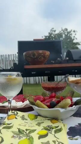 The video begins with a close-up of a table set for an outdoor meal. A glass of sparkling water with lemon and ice sits prominently in the foreground. Behind it, a platter with white cheese and slices of meat, likely salami, is visible. Further back, a bowl of what appears to be a tomato-based salsa or sauce is centered on the table, next to a small bowl filled with olives, pickles, and cherry tomatoes. A white bowl with a traditional folk pattern sits on a black grill, which is actively smoking, indicating food is being cooked. Next to the grill is a wooden table with a glass mug containing a light-colored beverage, possibly beer, and a brown bottle of beer. To the right of the grill, a glass dish overflowing with cooked shrimp is displayed. The scene transitions to focus on the grill, where skewers of meat are sizzling and producing smoke. The background consists of a grassy yard, a white picket fence, and trees under an overcast sky. The camera pans slightly to show more of the grill and the surrounding picnic area.